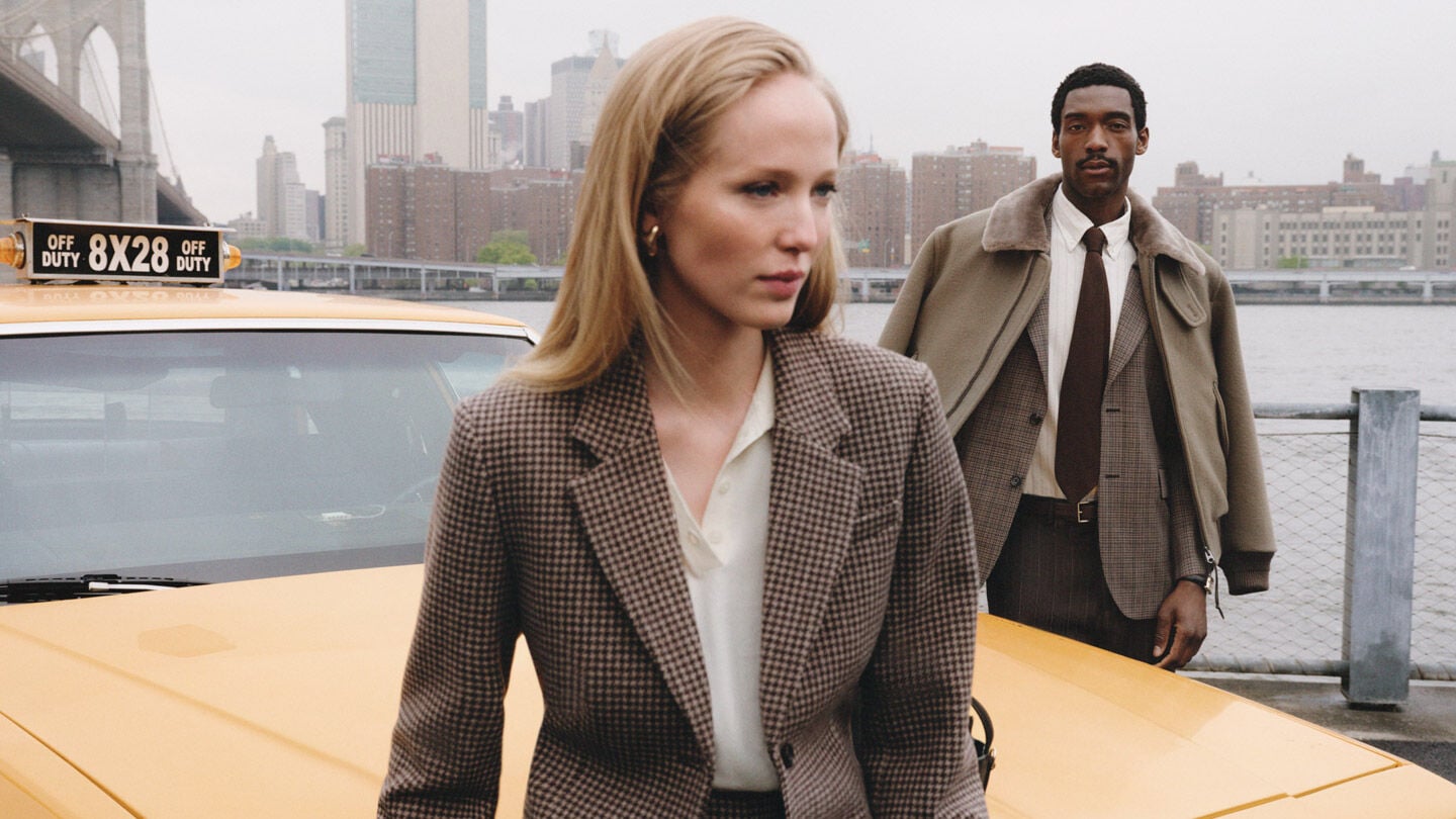 Two models standing next to a yellow taxi at a waterfront setting, woman styled in a Gant checked blazer and man styled in a Gant tailored jacket over a checked blazer.